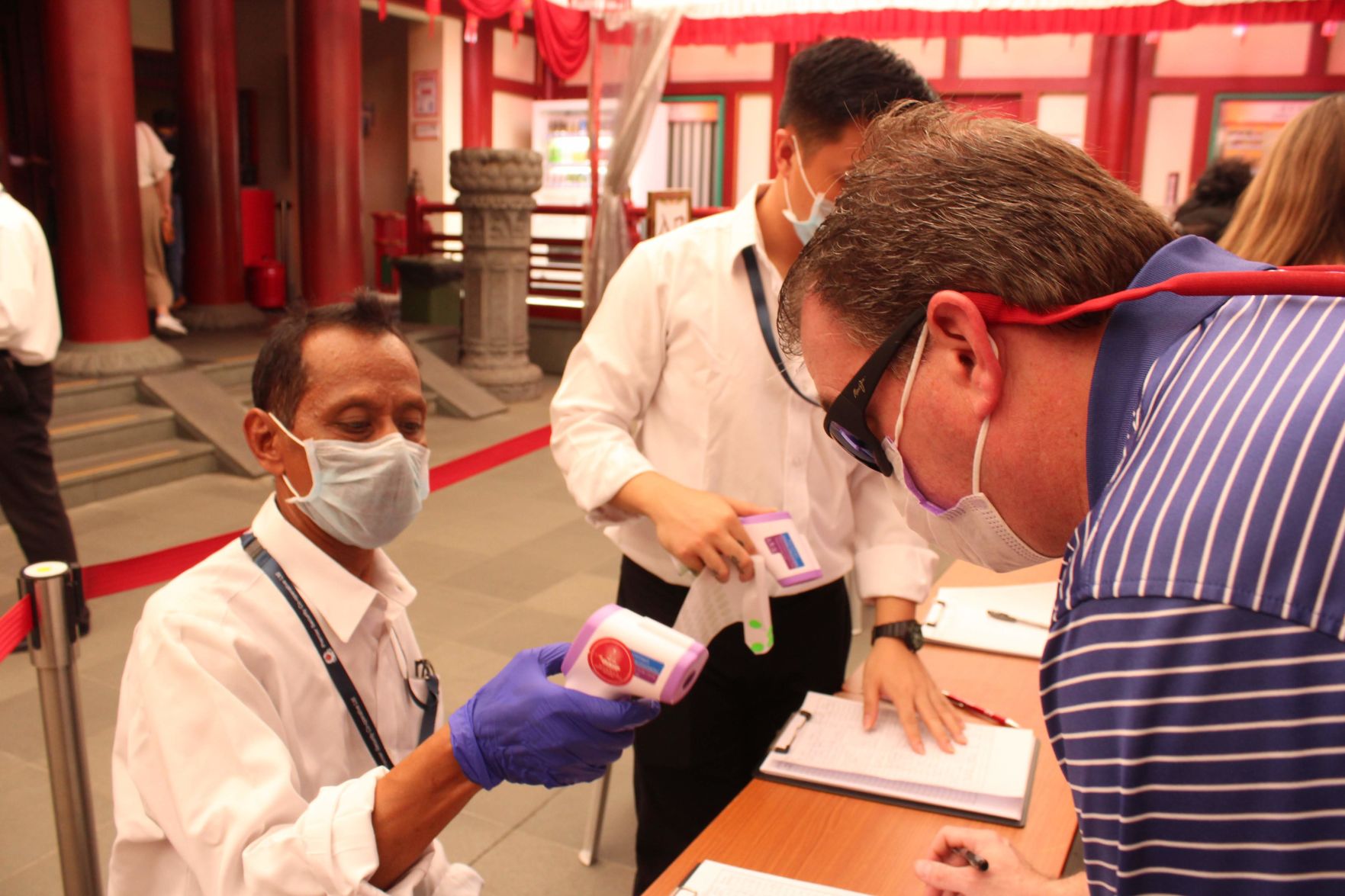A man in a mask holds a thermometer toward another man in a mask who leans closer to the desk
